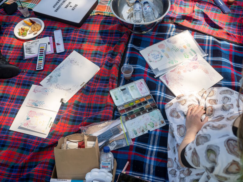 A overlay shot of watercolour materials and food laid on a picnic blanket